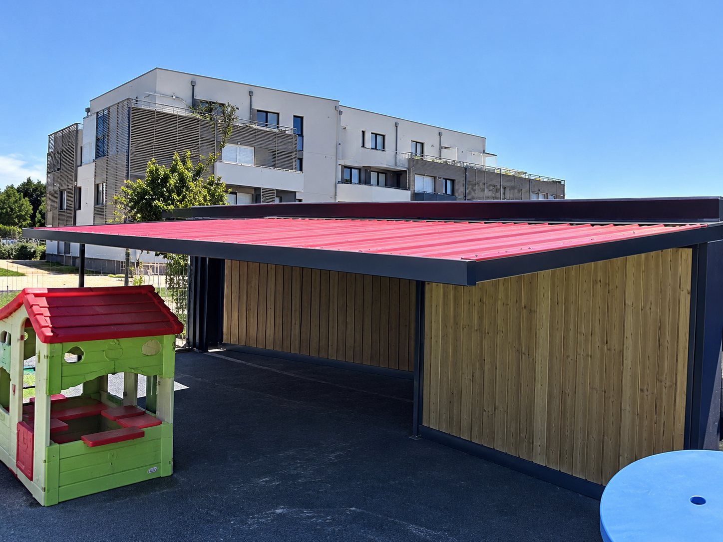 Un grand abri à toit rouge et parois en bois se trouve sur une pelouse, devant un bâtiment moderne et sous un ciel bleu.