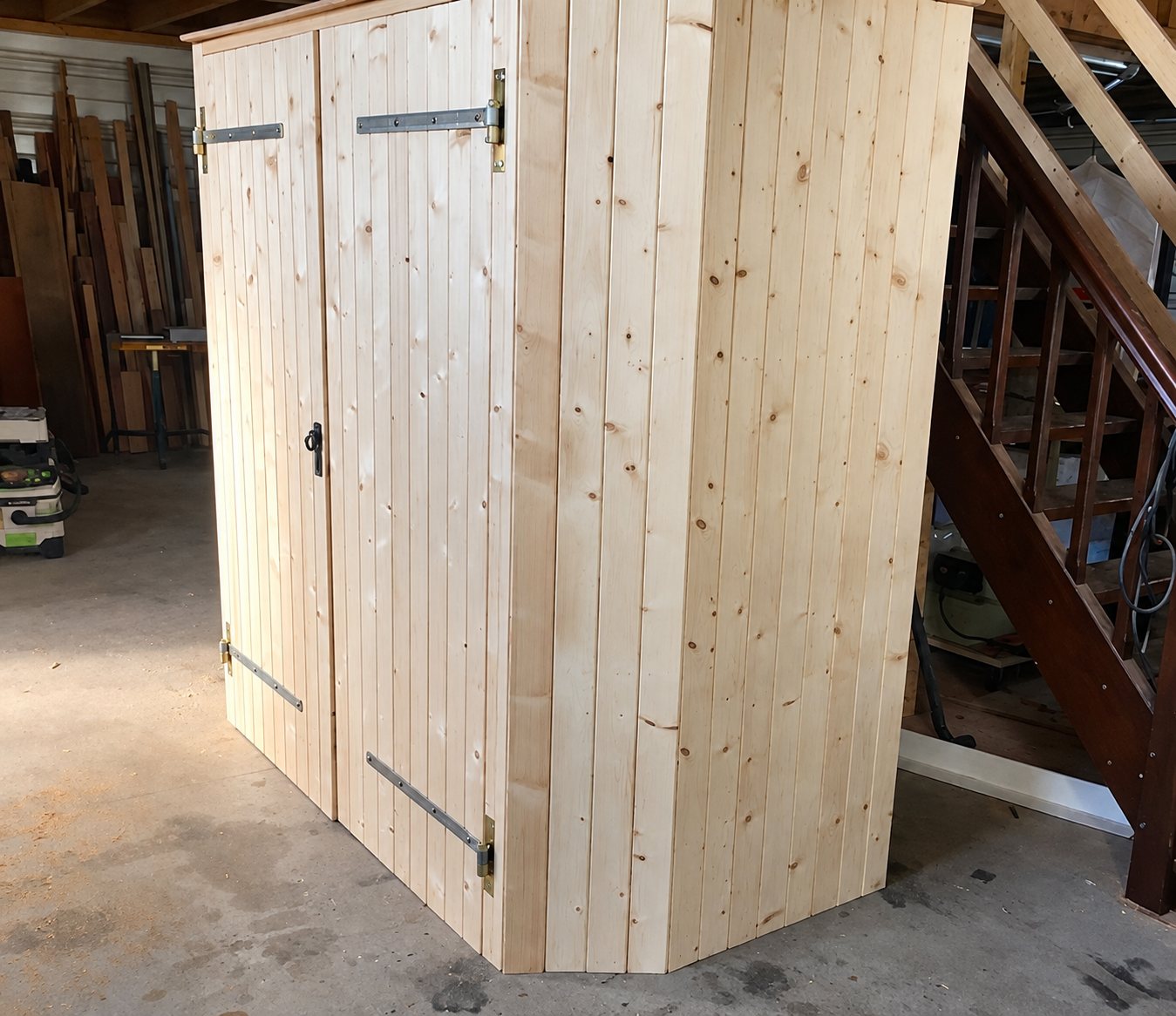 Une armoire en lames de sapin clair, fermée par deux portes à pentures métalliques, est installée dans un atelier à côté d'un escalier en bois.