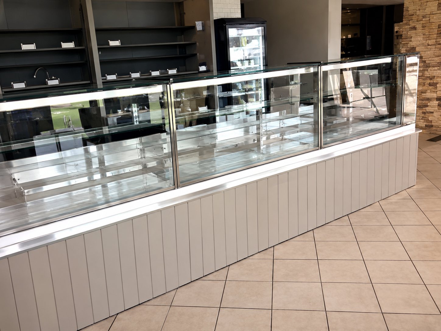 Un long comptoir vitré avec façade grise et étagères vides à l'intérieur d'un commerce, sur un sol carrelé clair.