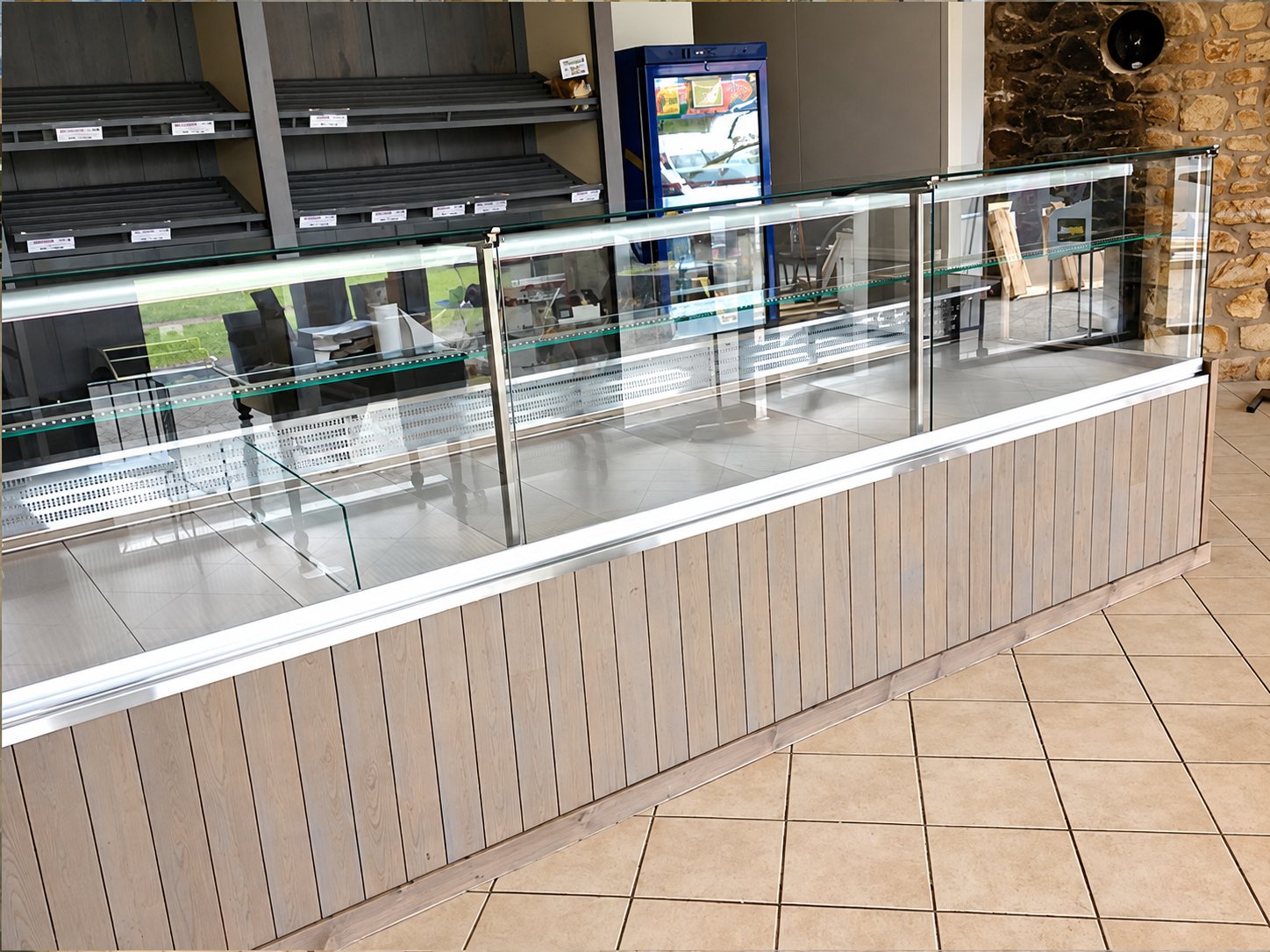 Un long comptoir réfrigéré en verre à façade de lattes de bois clair, vide, devant des étagères vides et un réfrigérateur bleu dans un commerce.
