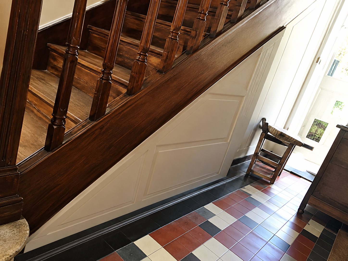 Un escalier en bois sombre avec une porte de placard blanche sous les marches, au-dessus d'un sol en carreaux colorés.