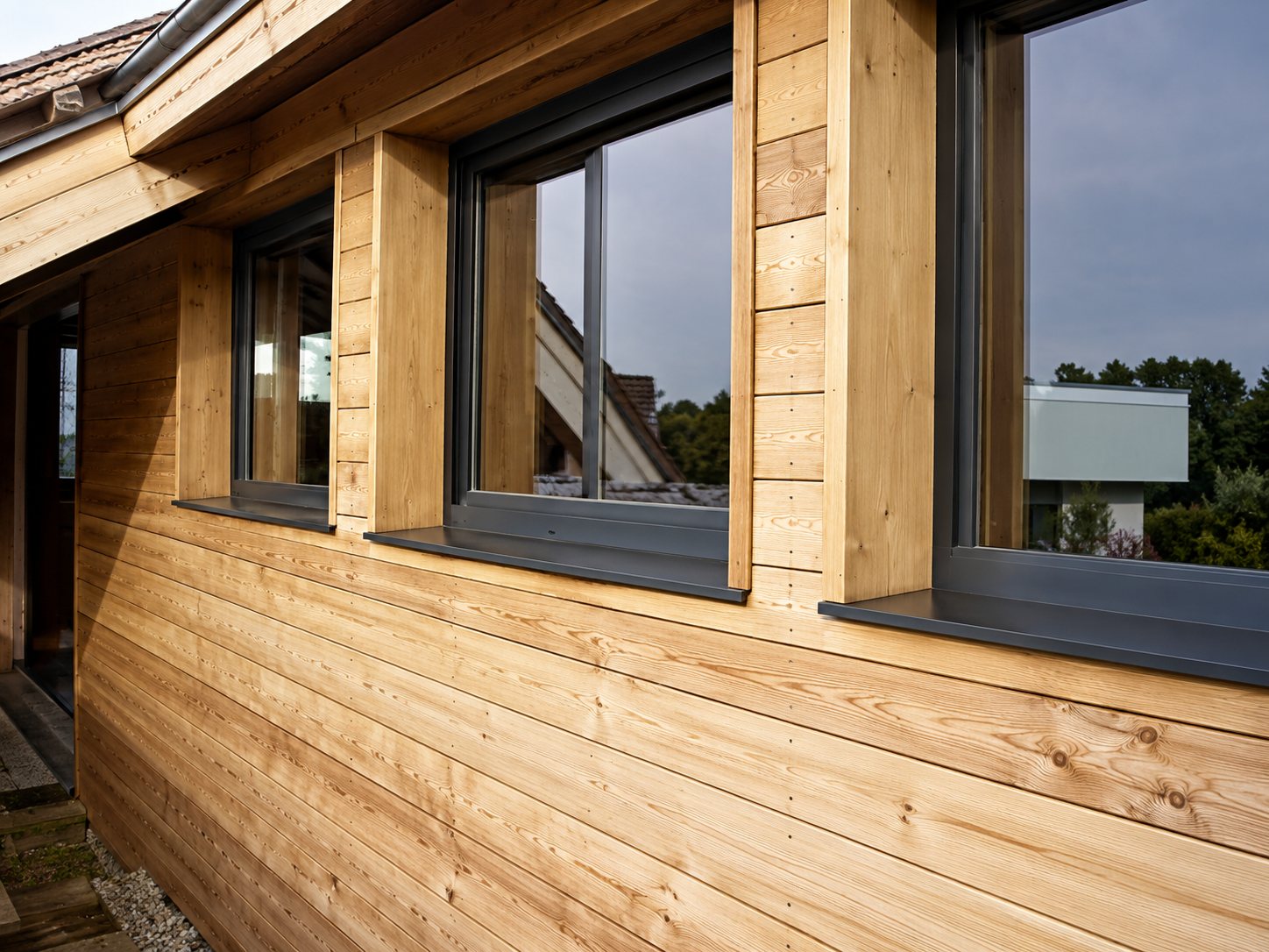 Façade d'un bâtiment recouverte de bardage en bois clair, percée de grandes fenêtres reflétant le ciel.