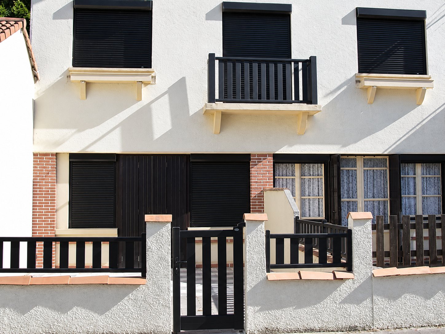 Façade d'une maison crépie avec volets roulants sombres, un balcon en bois et un portillon avec clôture ajourée au premier plan.