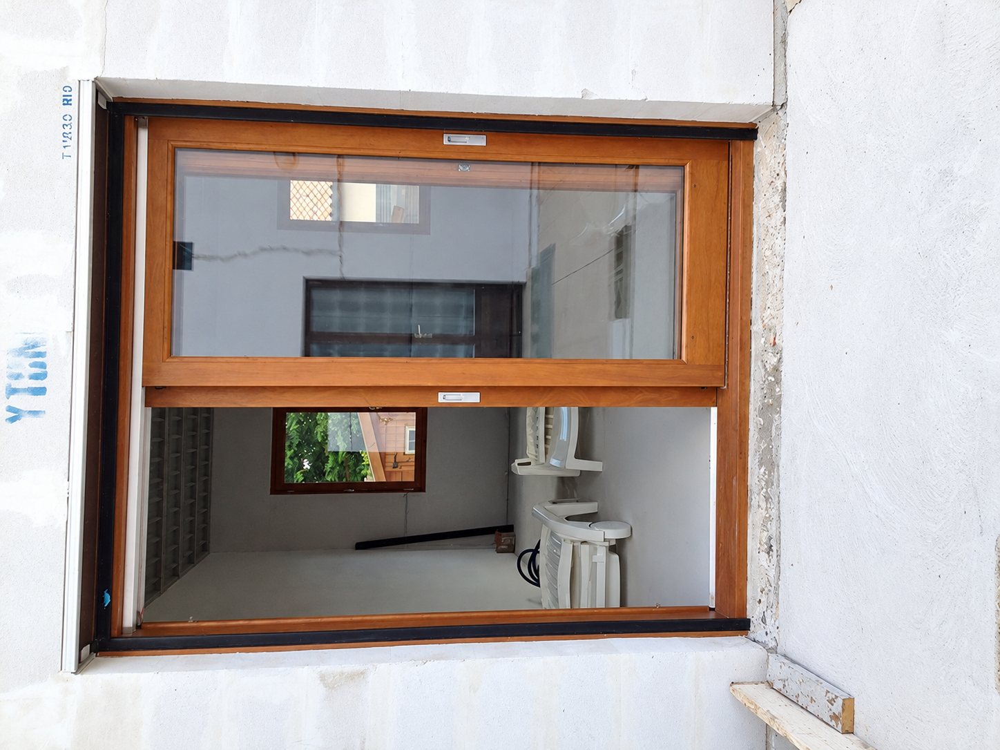 Une fenêtre coulissante à cadre en bois couleur chêne est posée dans un mur blanc, avec une pièce vide et des chaises en plastique blanches visibles à travers la vitre.