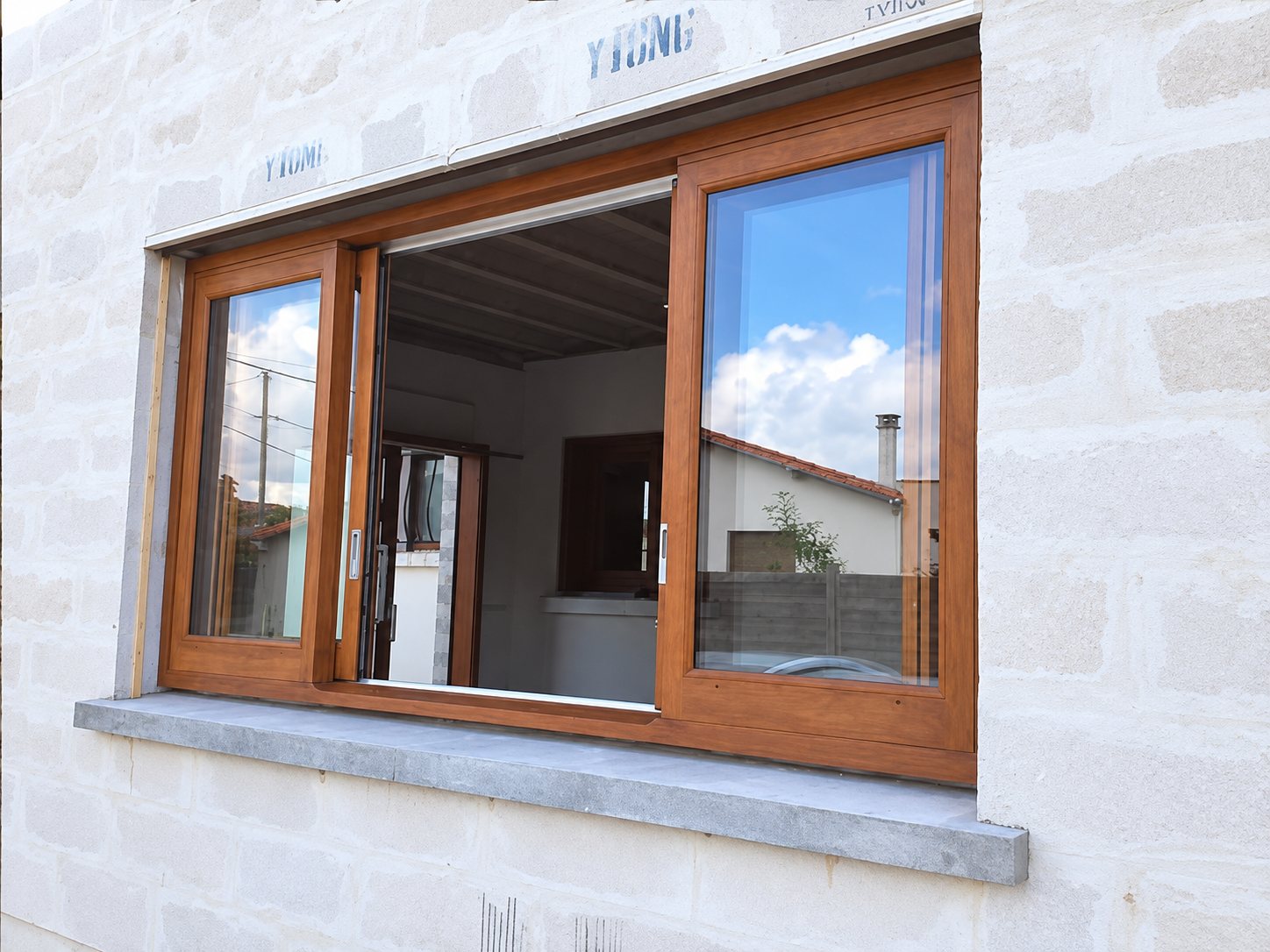 Fenêtre coulissante à cadre imitation bois posée dans un mur de blocs clairs d'une maison en construction, avec une tablette en pierre grise.