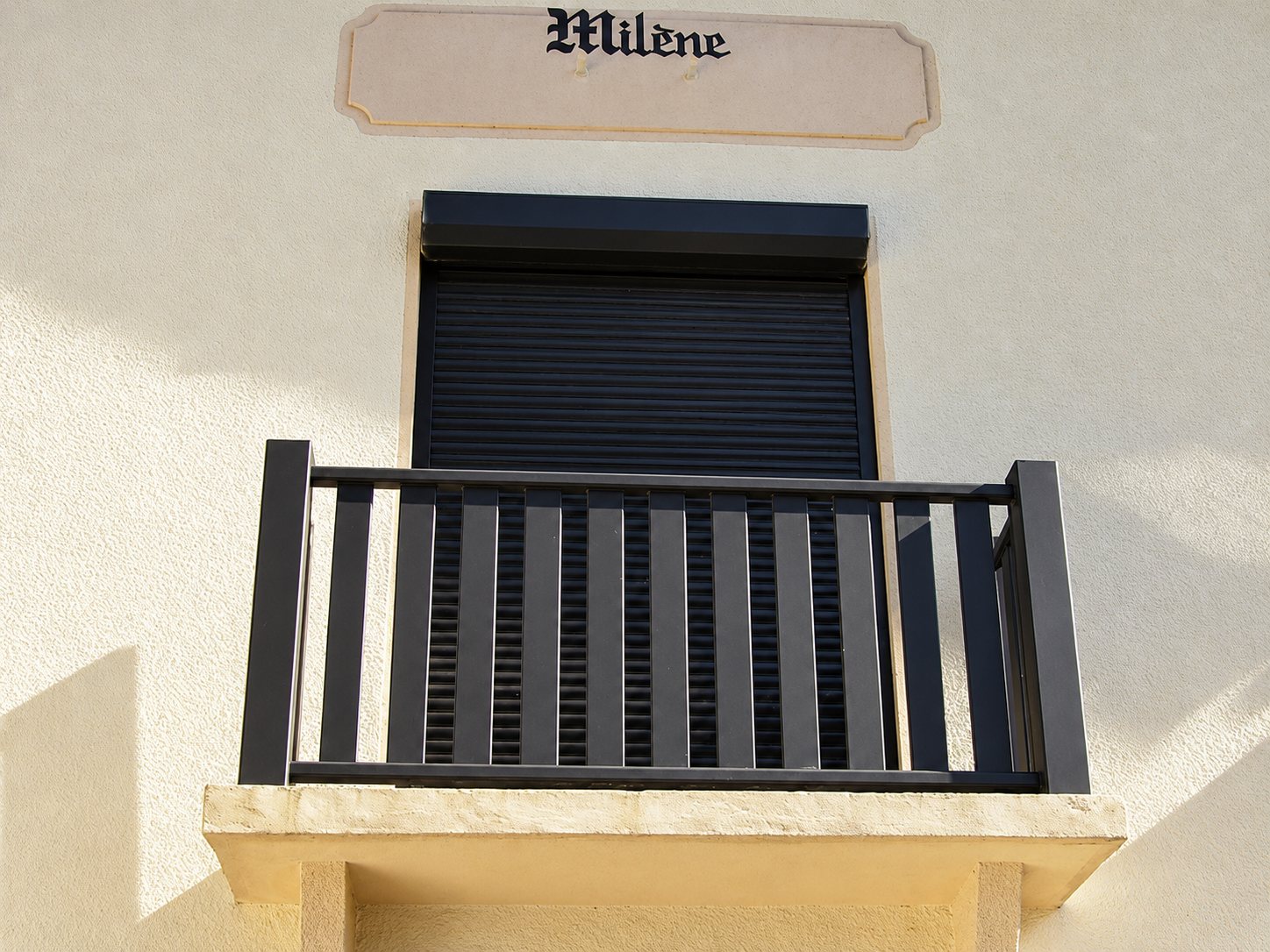Un balcon à garde-corps en lames brunes verticales devant une fenêtre à volet roulant fermé, sur une façade crépie portant une plaque au nom Milène.