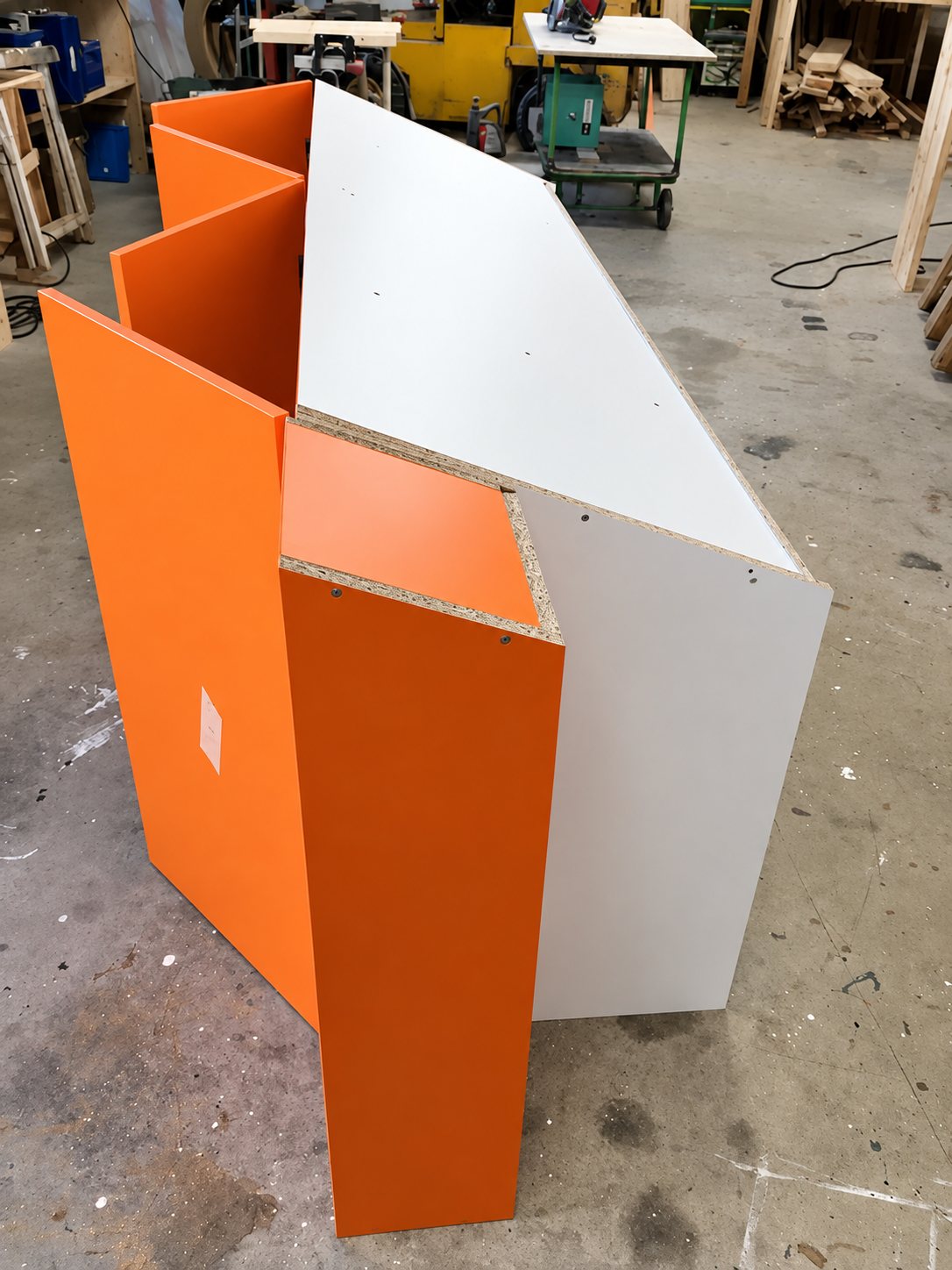 Un meuble de comptoir en panneaux de particules orange et gris, en cours d'assemblage sur le sol en béton d'un atelier de menuiserie.
