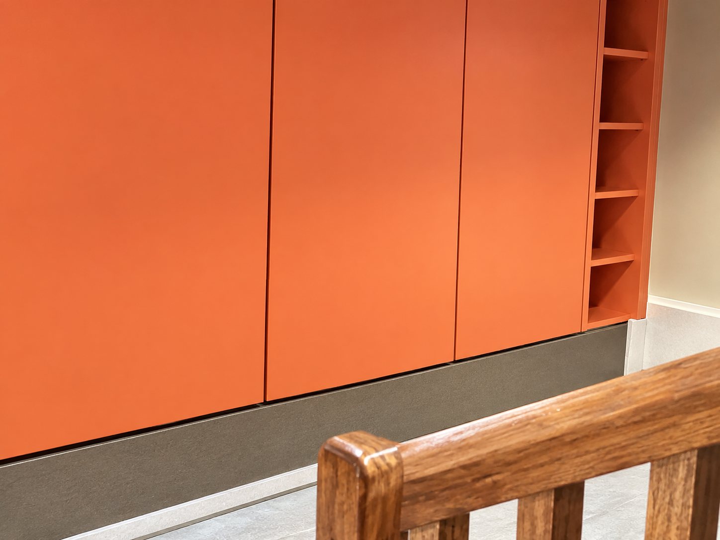 Un mur orange composé de grands panneaux lisses avec des étagères sur la droite, et une rampe en bois au premier plan.