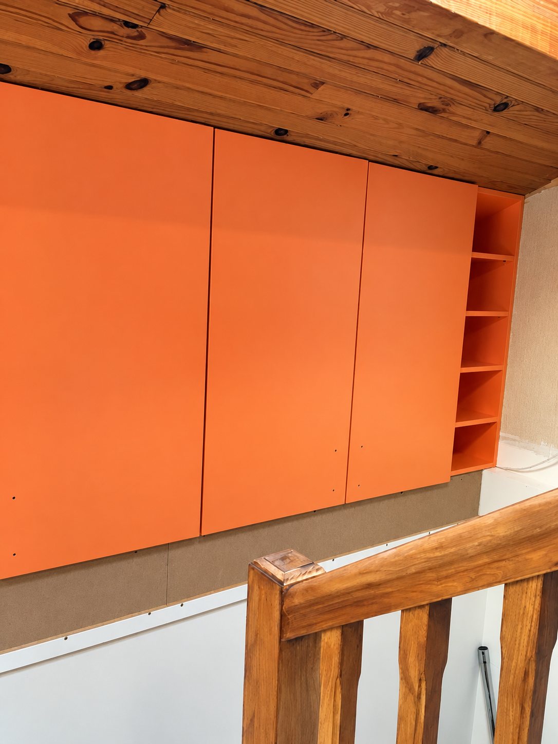 Un grand placard mural orange à trois portes avec étagères ouvertes sur le côté, installé sous un plafond en bois au-dessus d'une rampe d'escalier en bois.