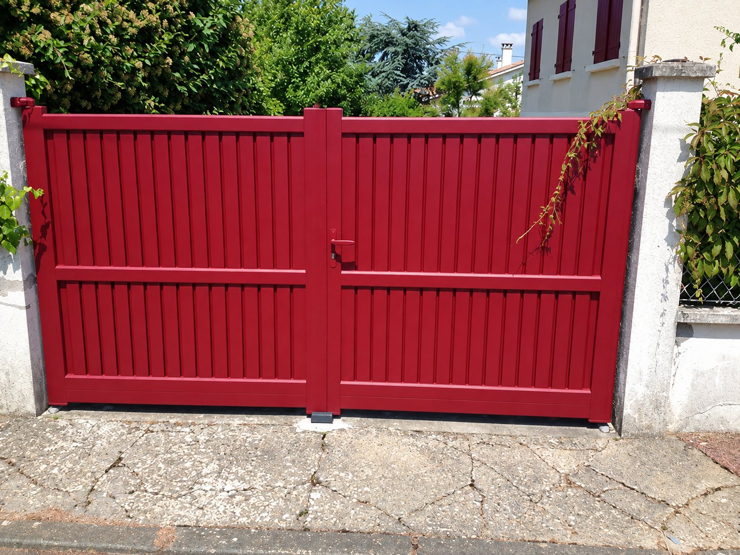 Un portail à deux battants rouge bordeaux à lames verticales, installé entre deux piliers blancs devant une maison et de la végétation.