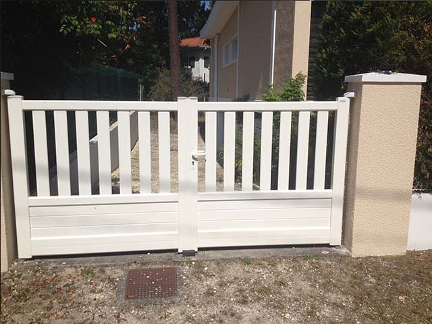 Un portail blanc à deux battants avec barreaux verticaux et partie basse pleine, installé entre des piliers crépis beige devant une maison.