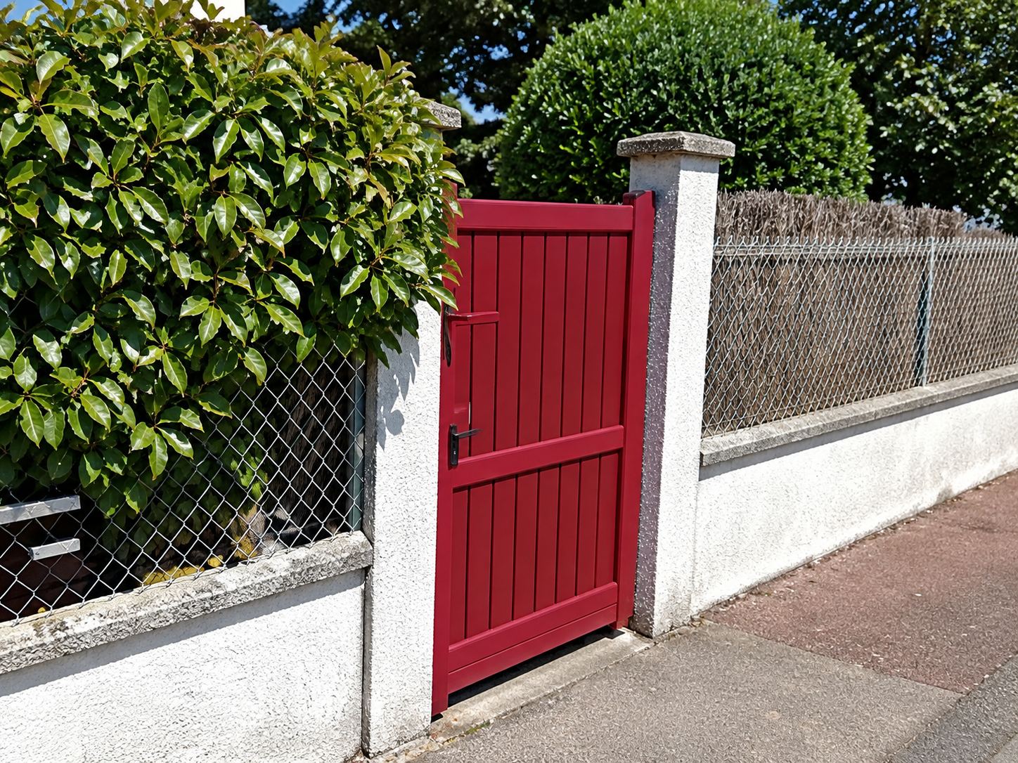 Un portail métallique rouge est installé dans une clôture à muret blanc, à côté d'une haie verte et d'un trottoir.