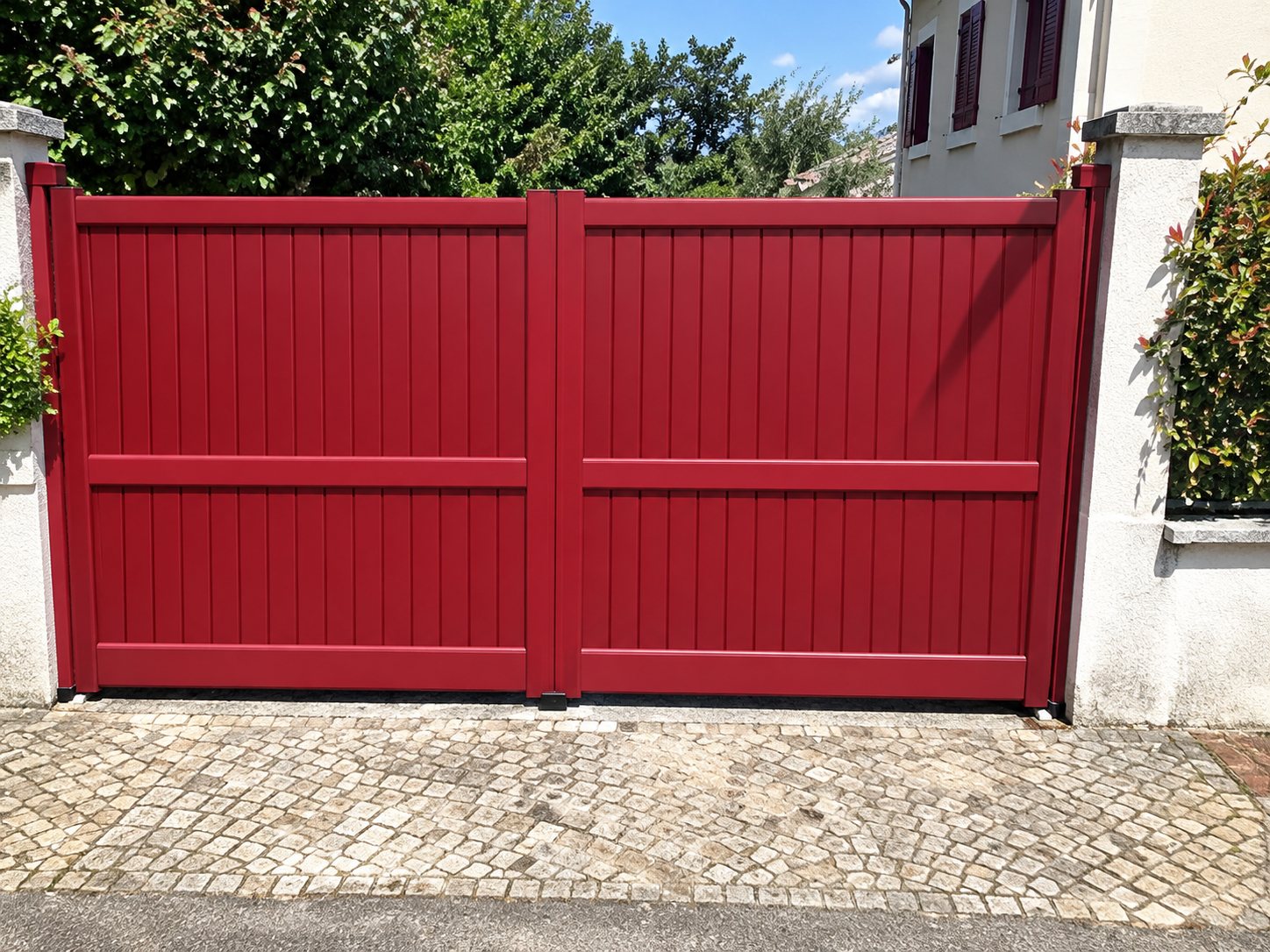 Un grand portail à lames pleines de couleur rouge bordeaux, fermé, encadré par des murets blancs devant une maison.