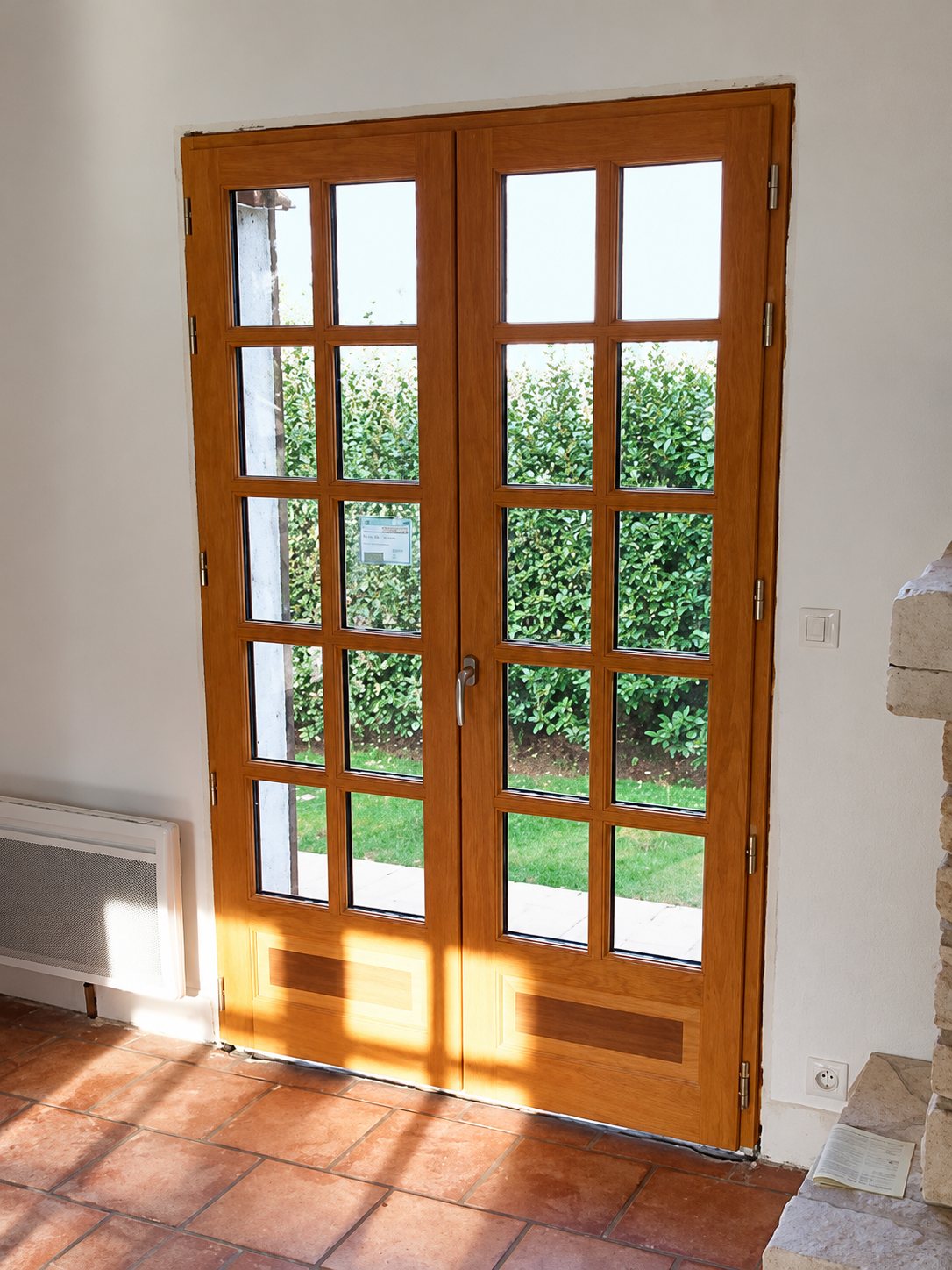 Une porte-fenêtre en bois à deux battants et petits carreaux, installée dans une pièce au sol en carrelage de terre cuite, ouvre sur un jardin avec une haie verte.