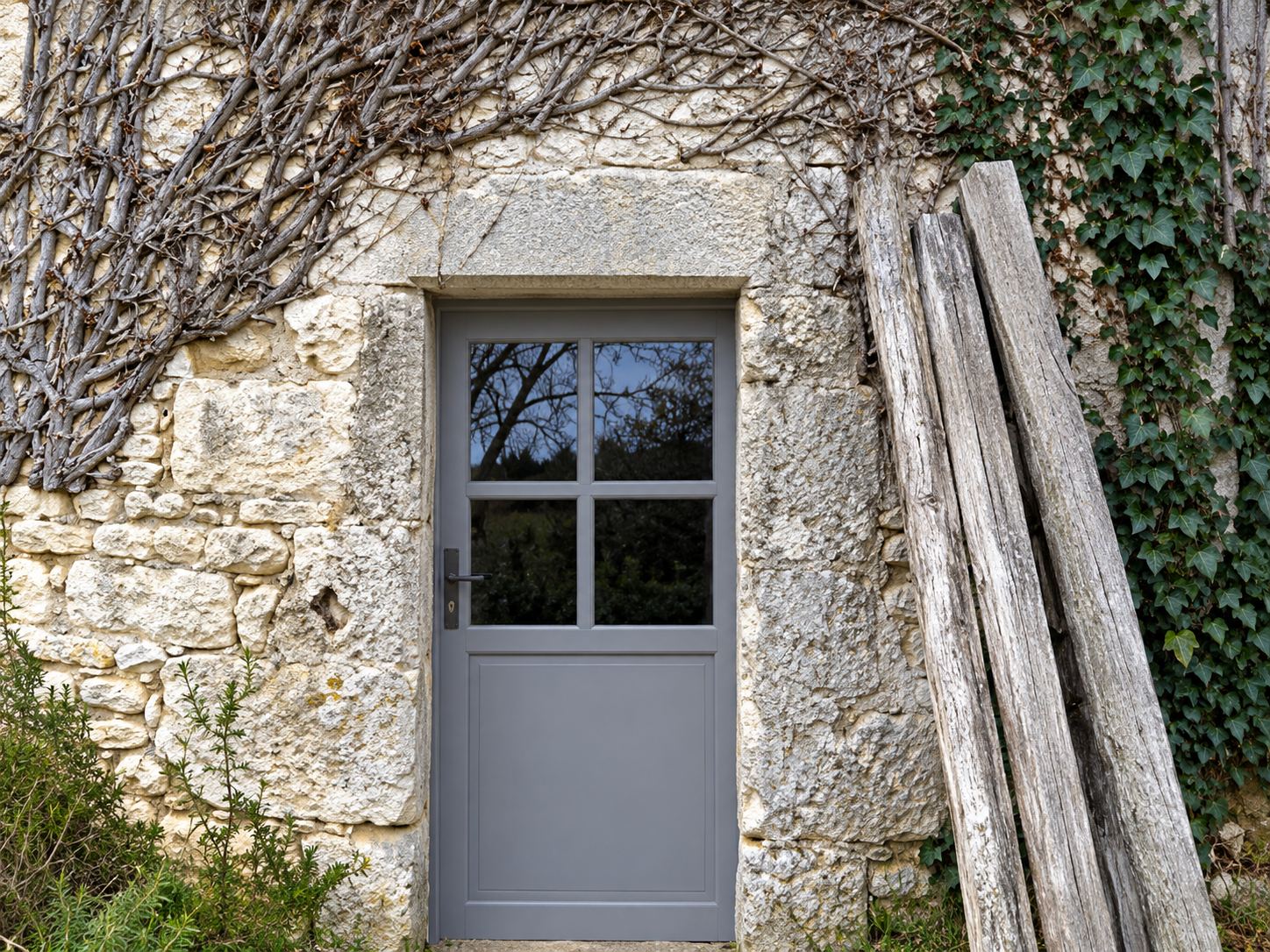 Une porte grise à petite fenêtre vitrée est encastrée dans un ancien mur de pierre en partie couvert de lierre, avec des planches appuyées à droite.