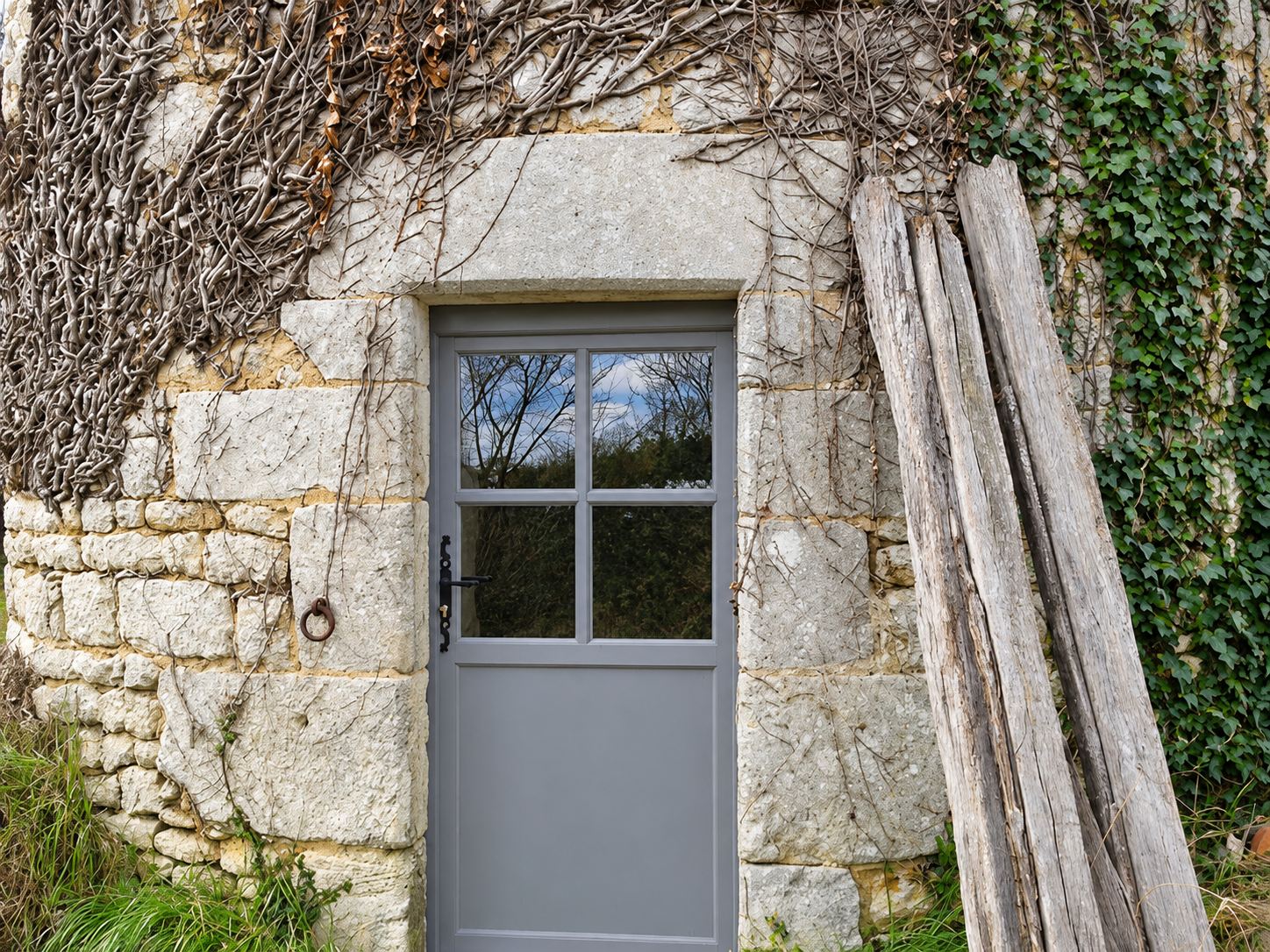 Une porte extérieure grise à petits carreaux est installée dans un mur en pierre ancien, avec du lierre et de vieilles planches en bois appuyées à droite.