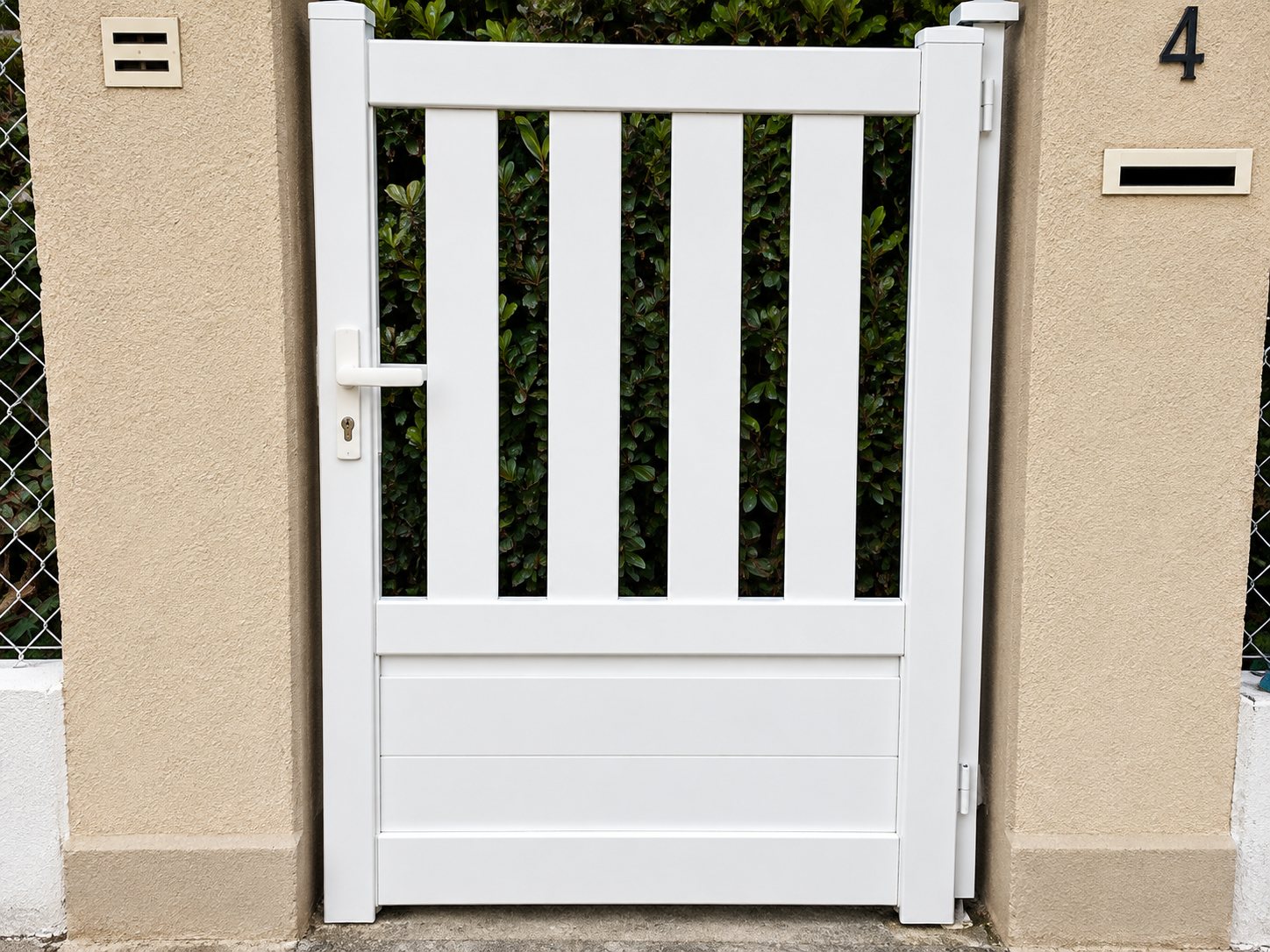 Un portillon blanc à barreaux verticaux est encadré par un mur beige, avec un numéro de maison visible à droite.