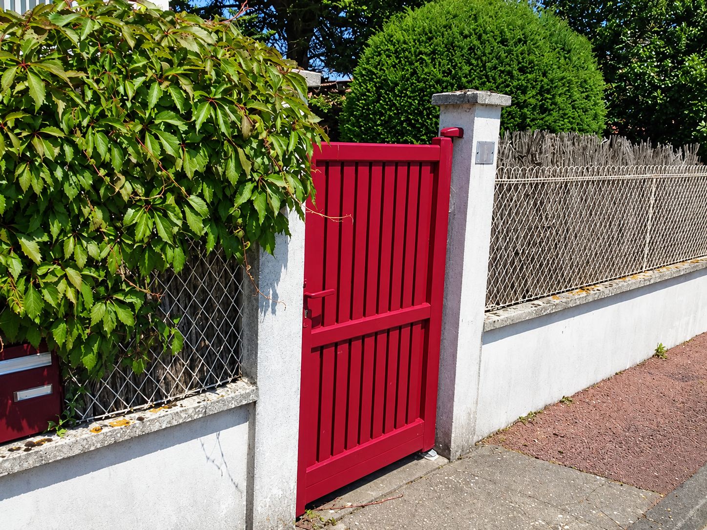 Un portillon en aluminium rouge à lames verticales est posé entre deux piliers en béton, au milieu d'une clôture grillagée et d'un muret blanc.