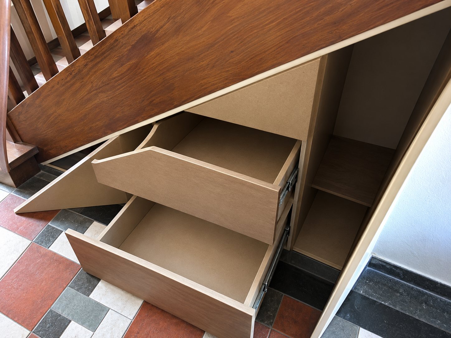 Rangement en bois clair aménagé sous un escalier, avec plusieurs tiroirs ouverts.