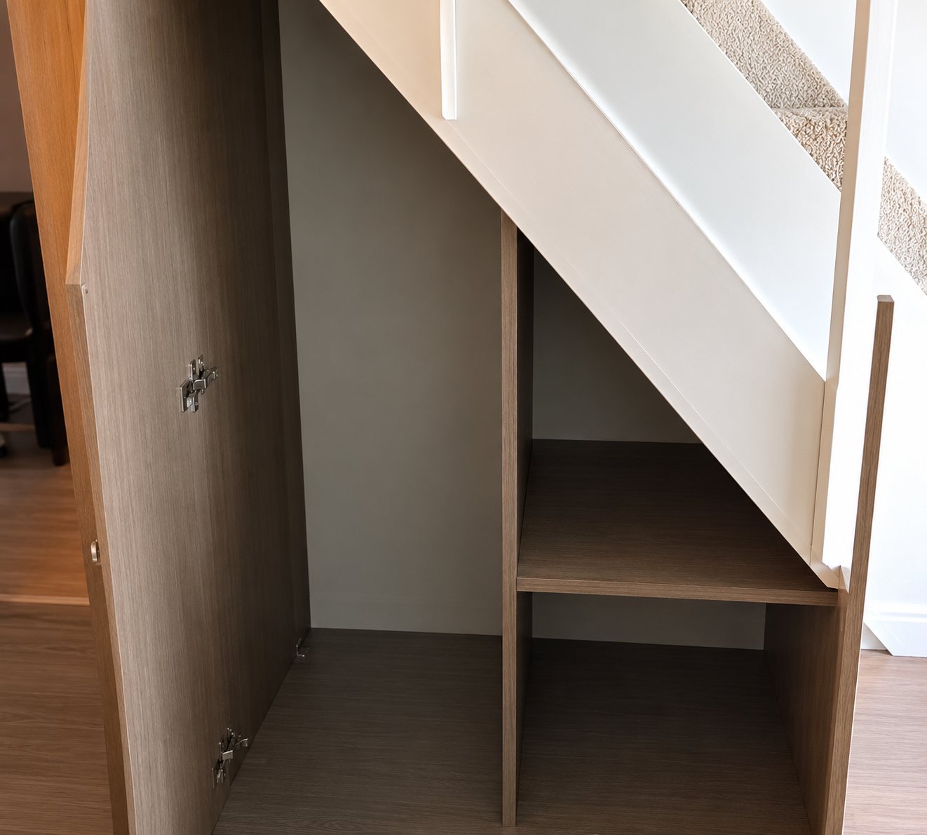 Placard avec étagères intégré sous un escalier en bois clair, portes ouvertes sur un intérieur sombre.