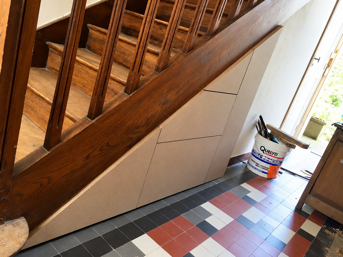 Des placards en panneaux bruts sont intégrés sous un escalier en bois foncé, au-dessus d'un carrelage ancien rouge, noir, gris et blanc, avec un seau de peinture près de la porte.