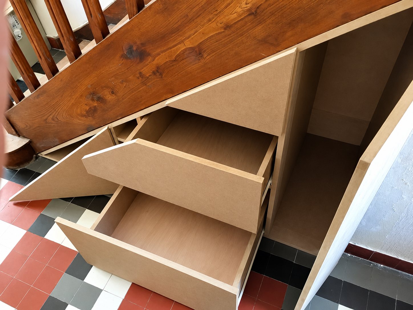 Tiroirs et placards en medium brut aménagés sous un escalier en bois, au-dessus d'un carrelage ancien à damier rouge, gris et blanc.