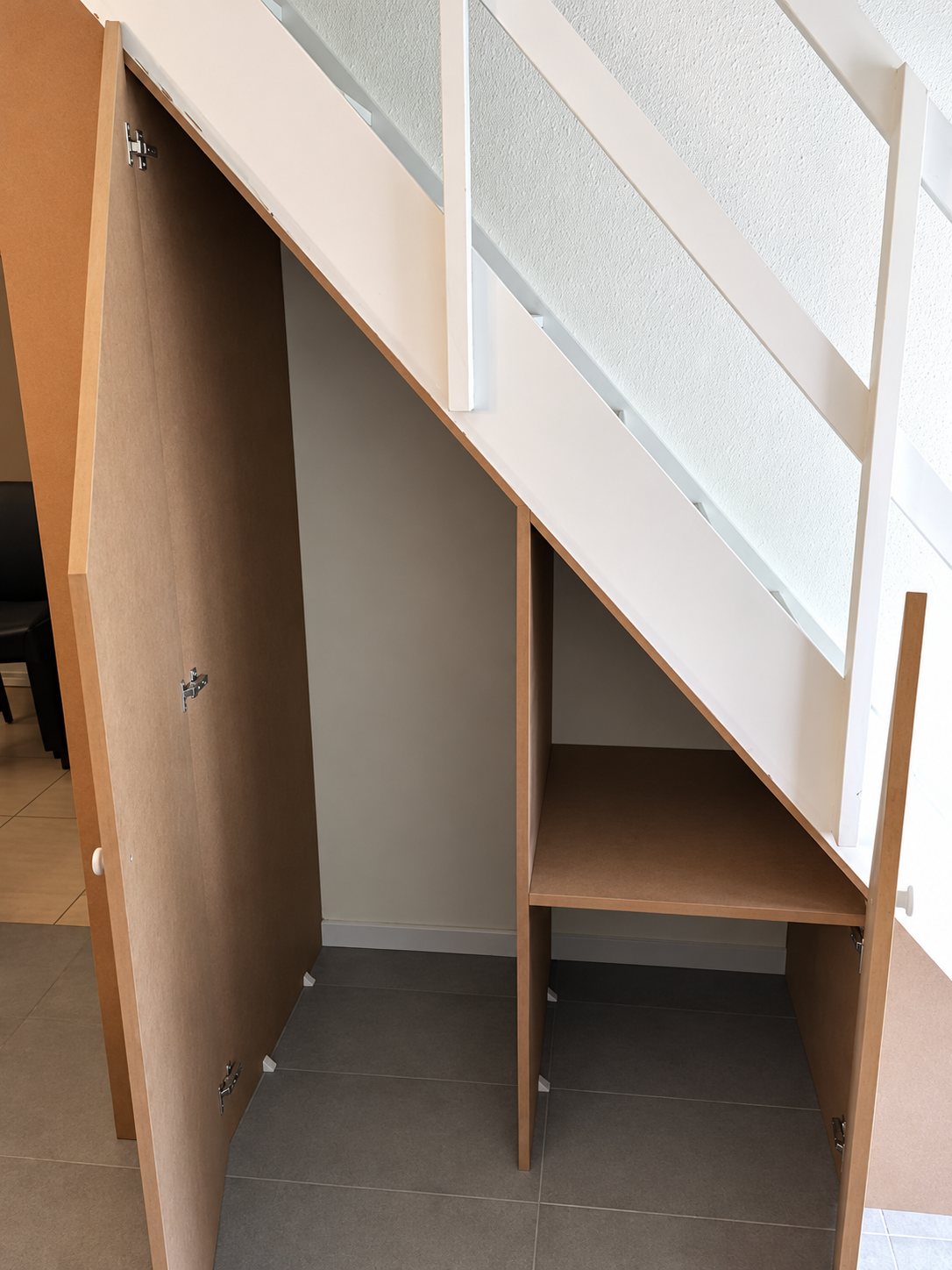 Placard sur mesure installé sous un escalier blanc, portes ouvertes, avec une étagère en bois et un sol en carrelage gris.