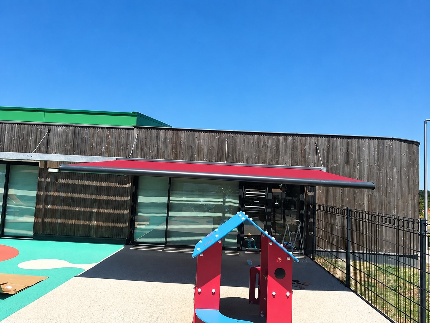 Un grand store banne rouge déployé sur la façade vitrée d'un bâtiment en bardage bois, à côté d'une aire de jeux colorée avec une petite maisonnette.