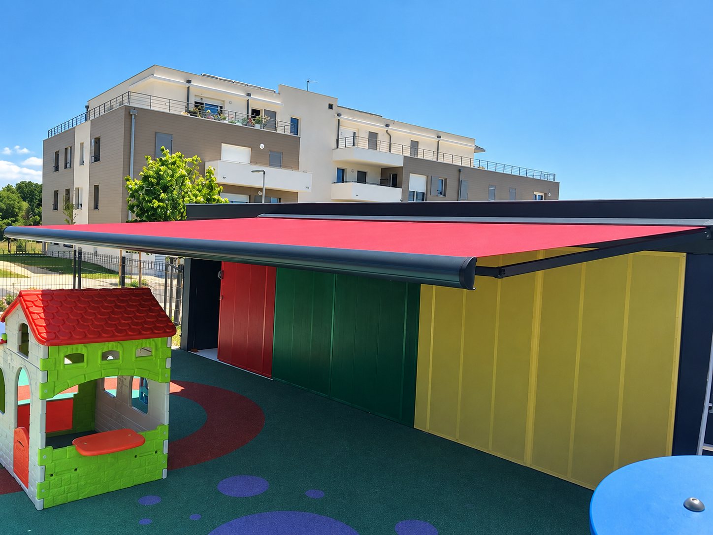 Un grand store banne rouge couvre un abri coloré dans une aire de jeux pour enfants, avec une maisonnette en plastique et un immeuble moderne à l'arrière-plan.