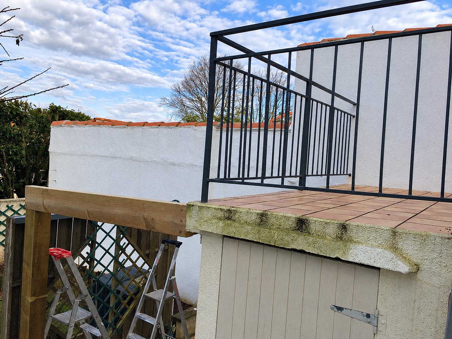 Une terrasse en carreaux de terre cuite avec un garde-corps métallique noir surplombe un petit local à porte en bois, près d'une poutre en bois neuve et de deux escabeaux.