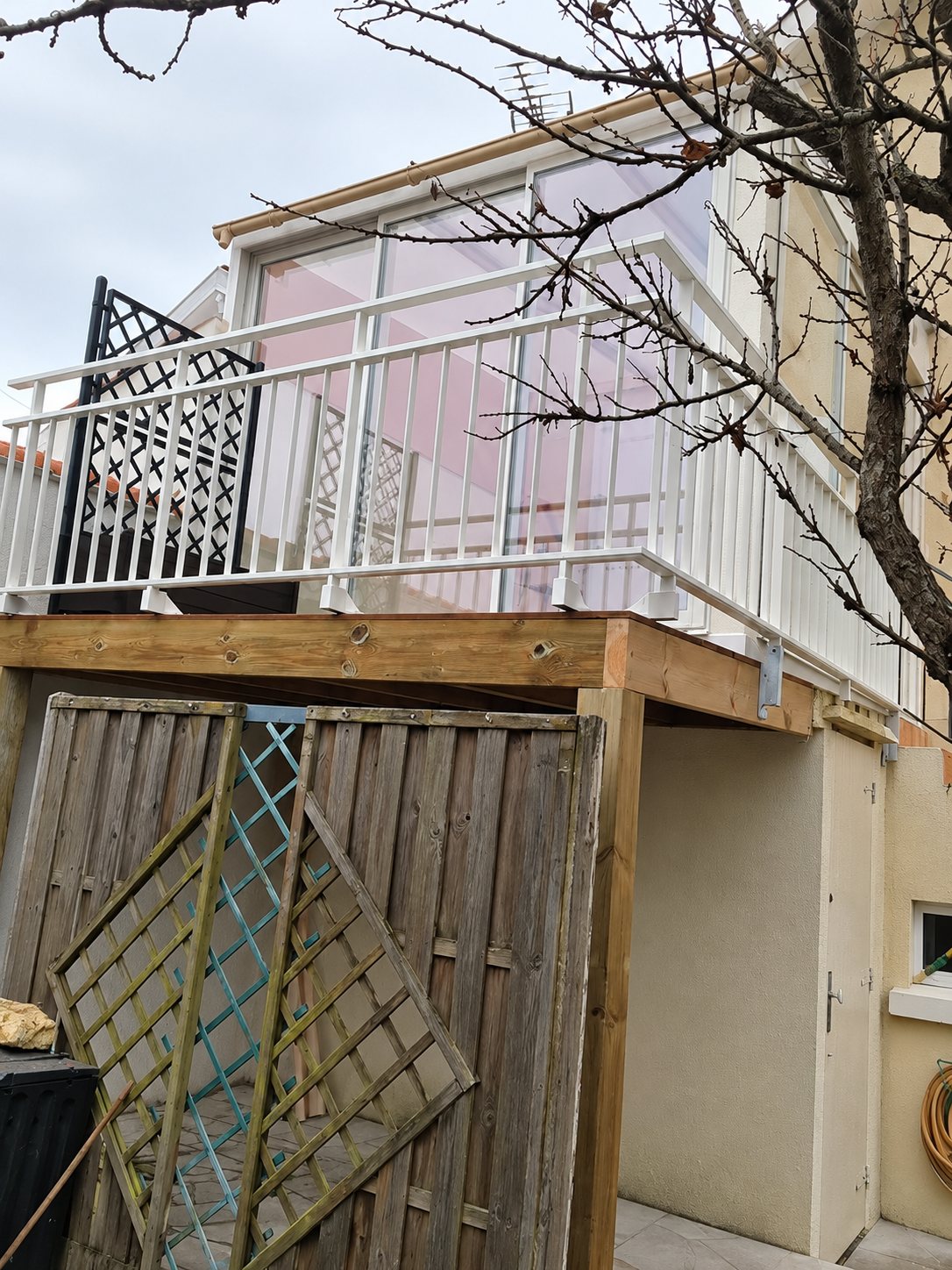 Terrasse surélevée à structure en bois équipée d'un garde-corps blanc devant une véranda, au-dessus d'un abri avec palissades en bois.
