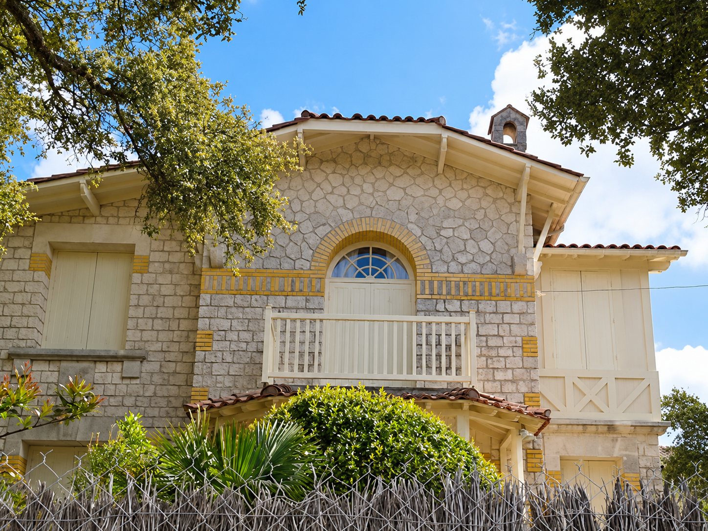 Façade d'une villa en pierre claire avec un balcon en bois blanc, des volets crème et un toit de tuiles, entourée d'arbres et d'une clôture.
