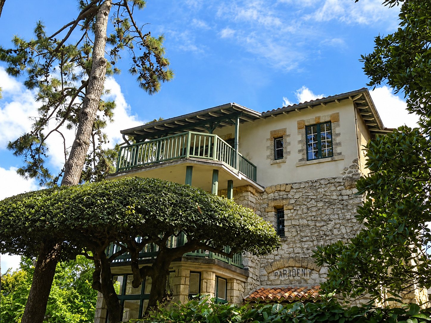 Une villa en pierre et enduit clair avec un balcon en bois vert, entourée de pins et d'arbustes sous un ciel bleu.